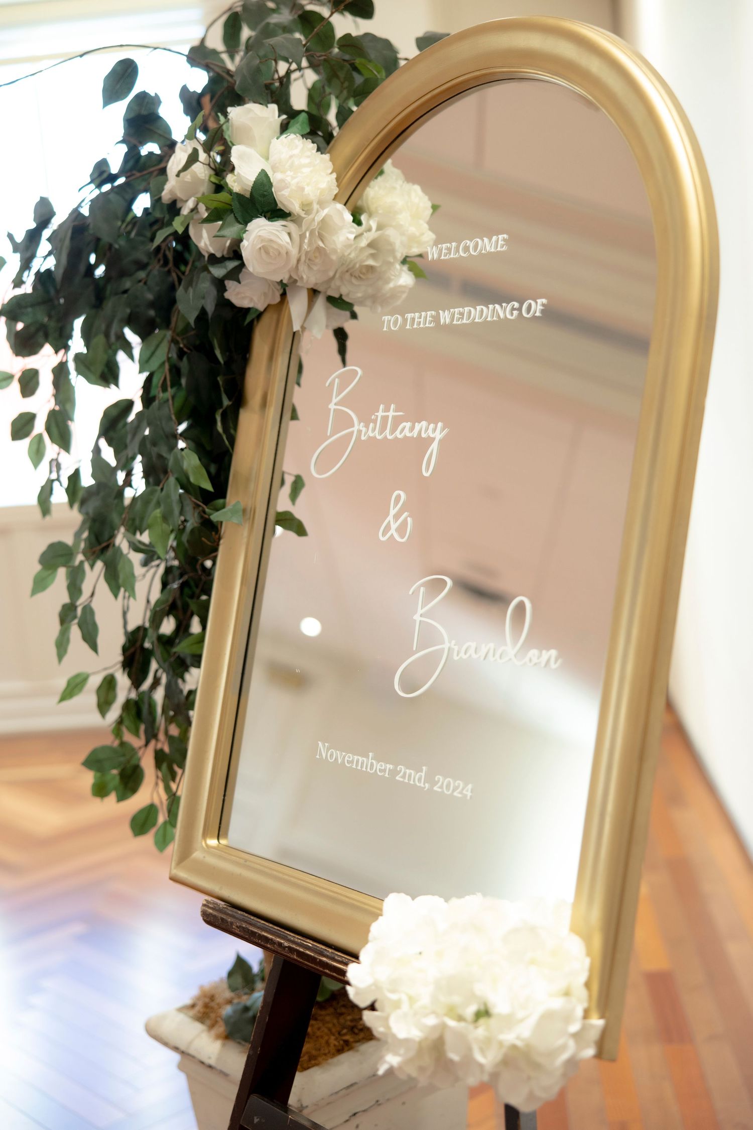 Elegant gold-framed wedding welcome sign decorated with white roses and greenery.
