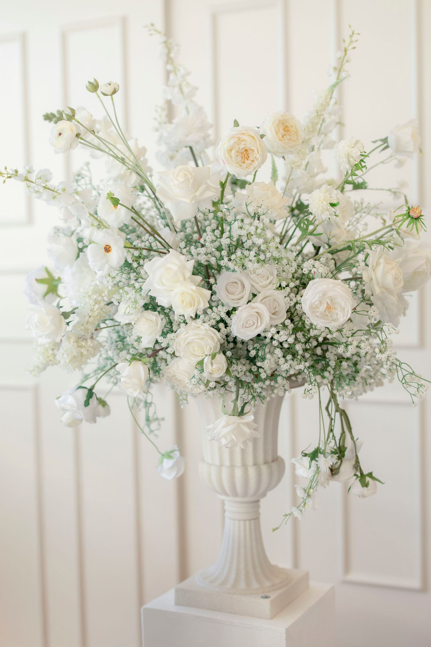 Elegant white floral arrangement with roses and baby's breath in a classic white pedestal vase.