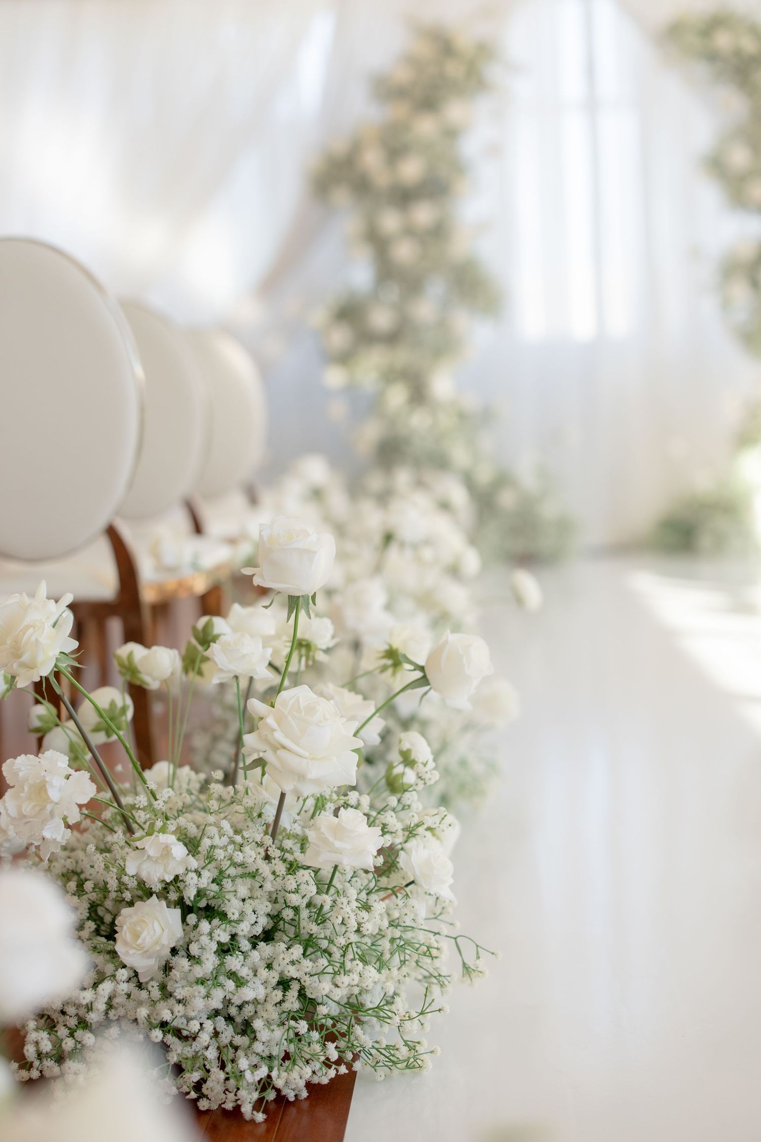 Delicate white roses and baby's breath floral arrangements line white ceremony chairs.