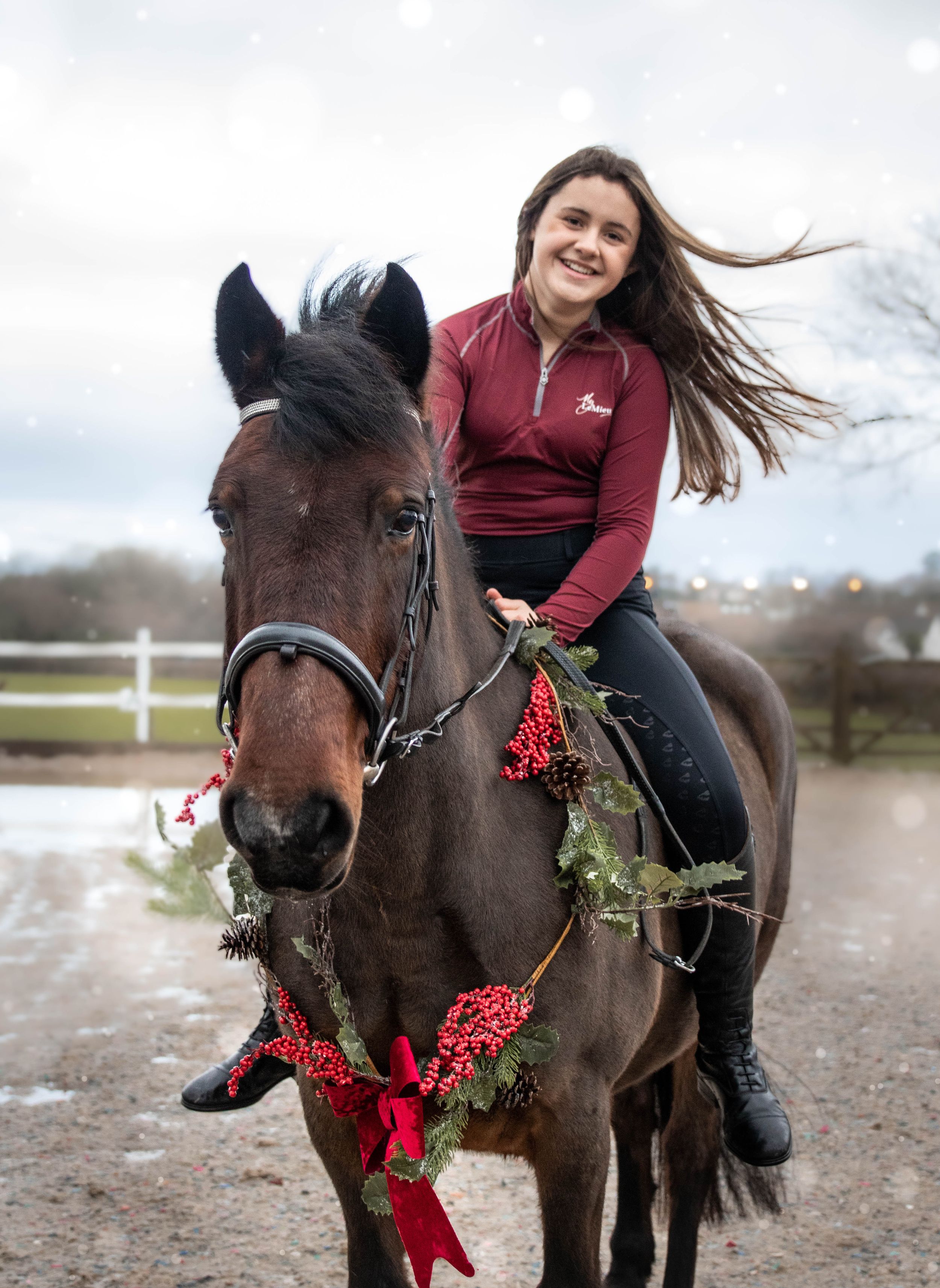 Pony adorned with Christmas decorations and red bow being ridden in an outdoor arena.