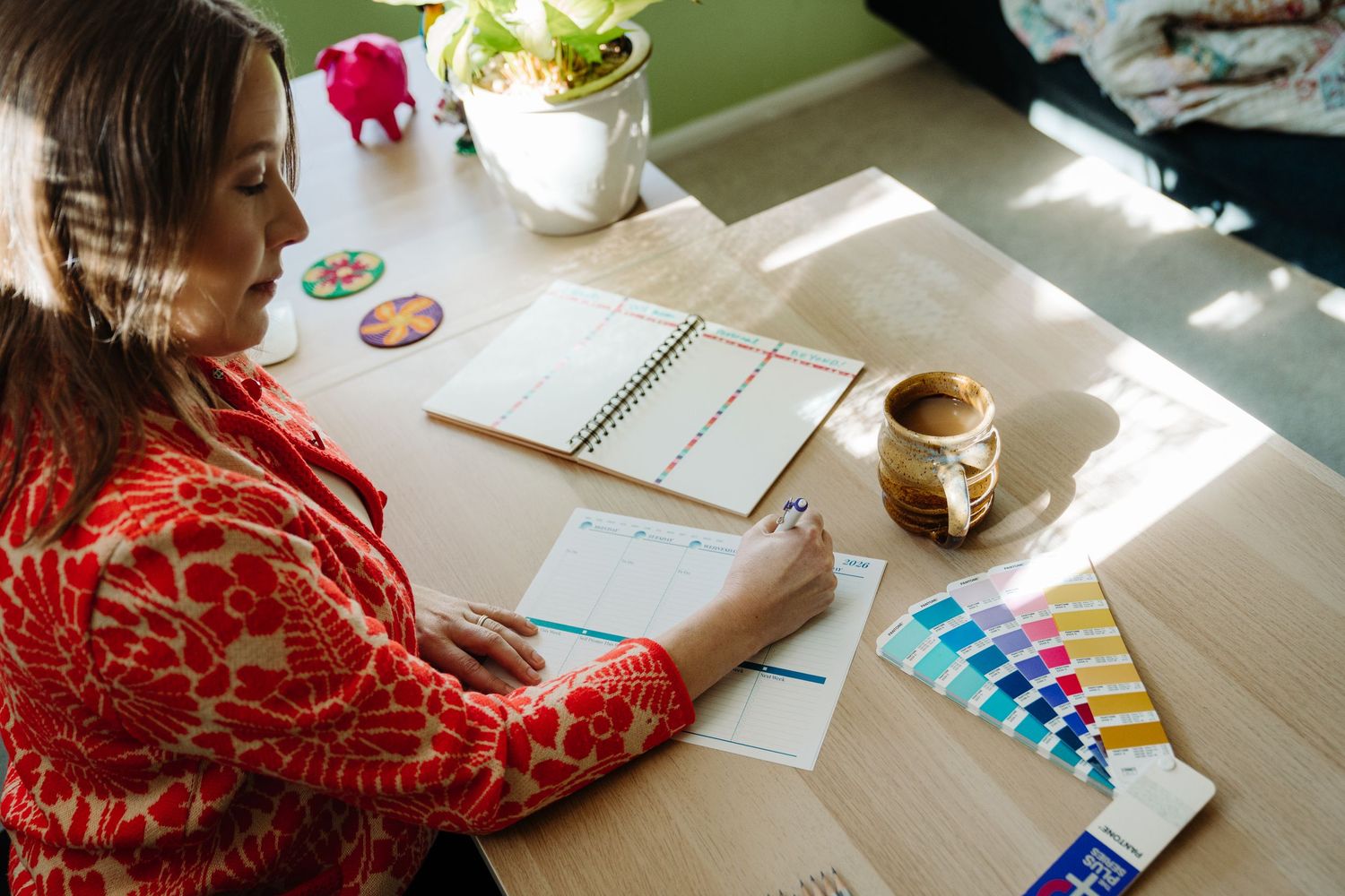 A person in a red sweater sits at a bright desk working on art with watercolor paints and paper.