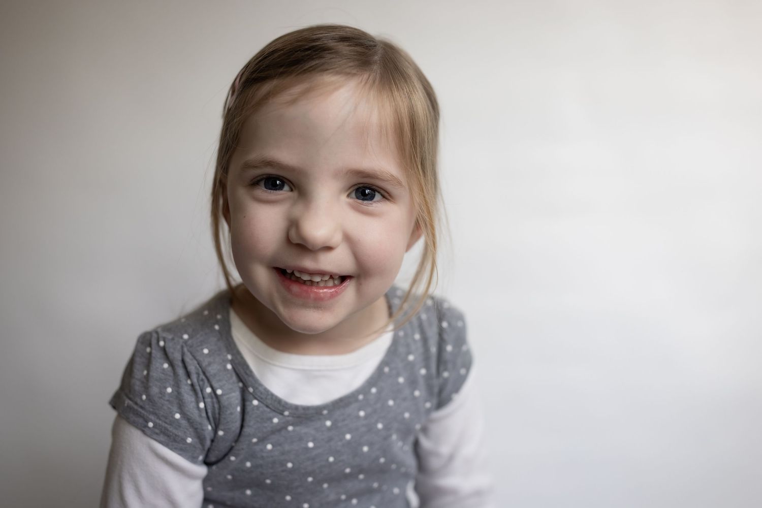 Young blonde girl in a gray polka dot shirt smiling brightly against a light gray background.