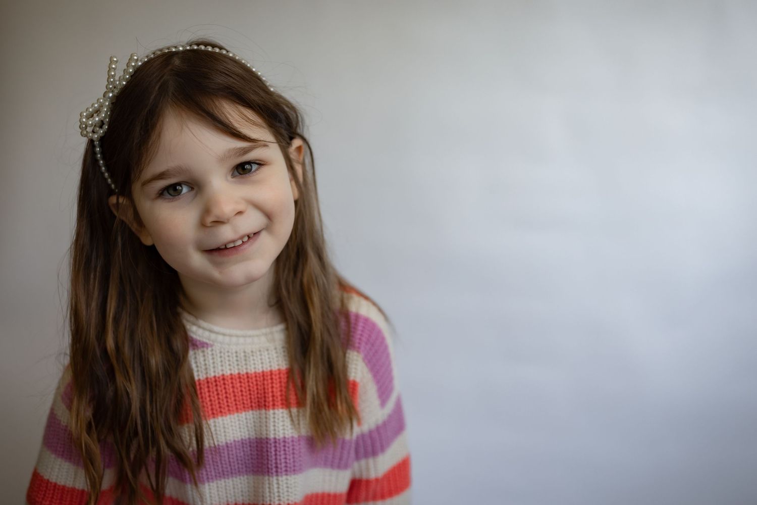 Sweet young girl with long brown hair wearing a pearl headband and pink striped sweater, smiling.