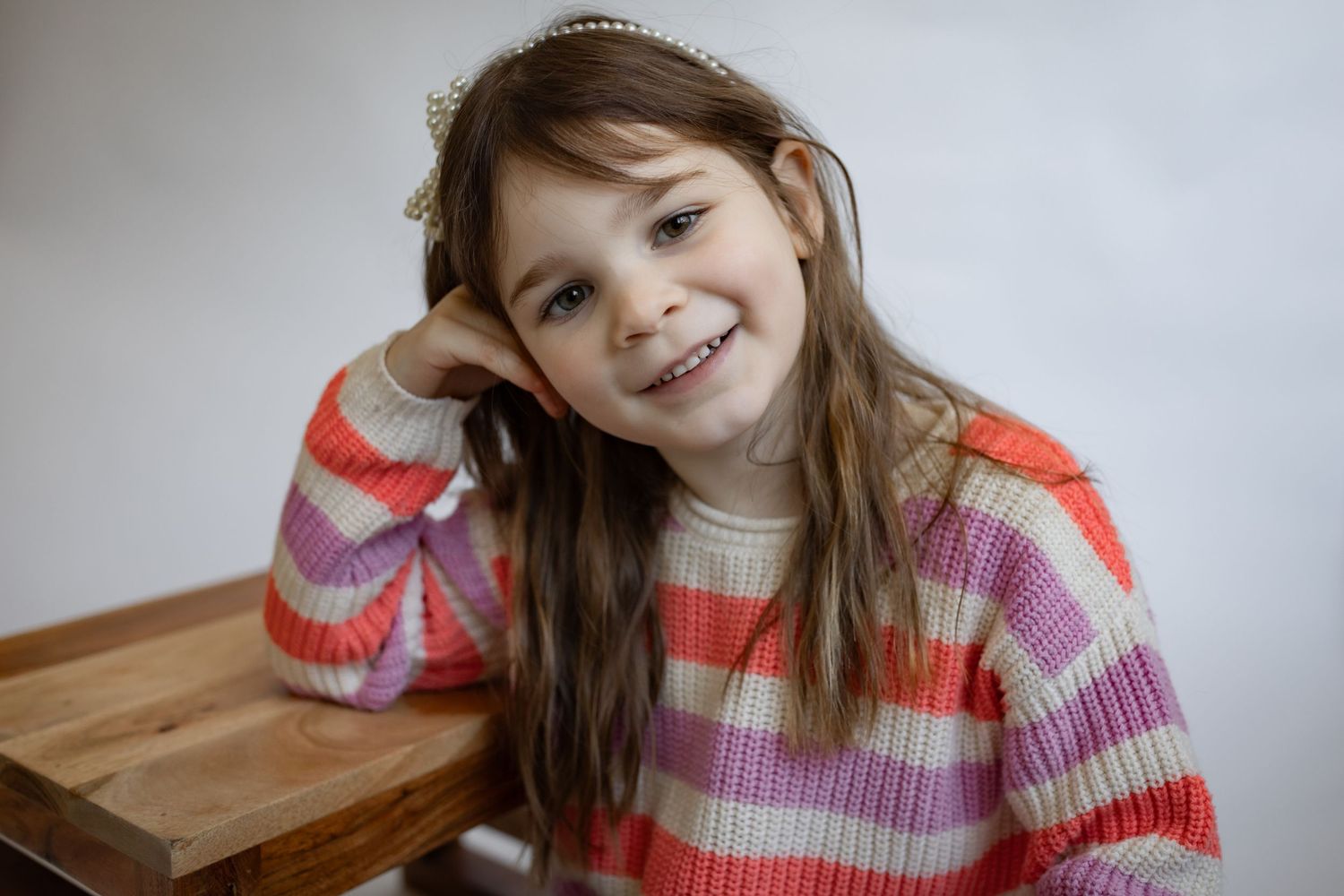 Young girl in a colorful striped sweater smiling while leaning on a wooden surface with a headband.