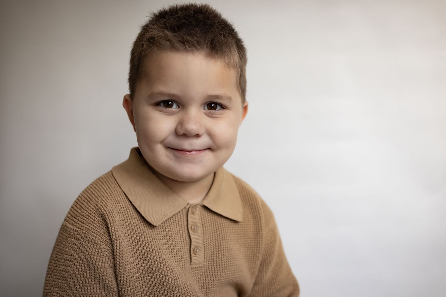 Smiling young boy with short brown hair wearing a tan polo shirt against a light gray background.