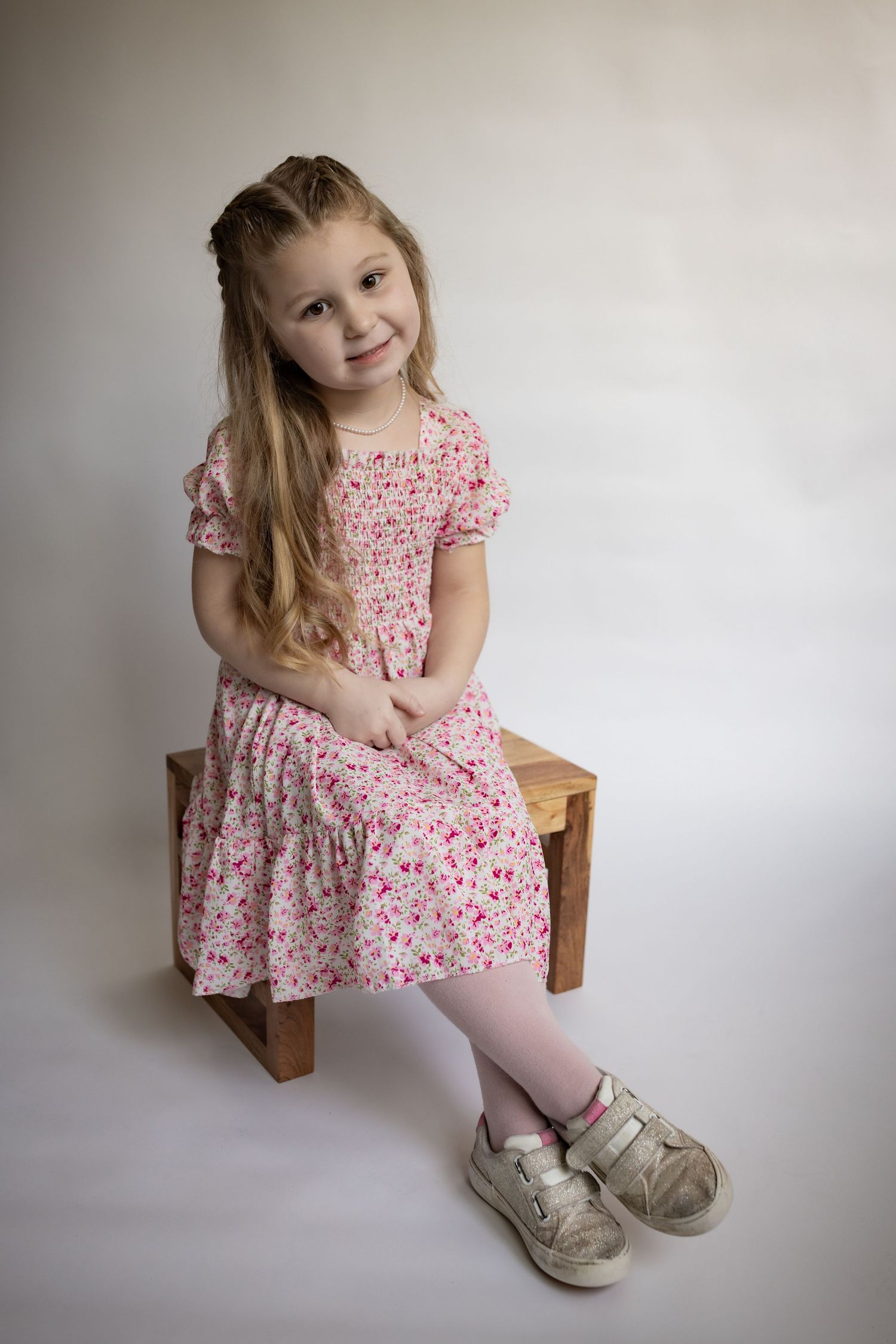 Young girl in a pink floral dress and pink tights sitting on a wooden stool wearing silver sneakers.