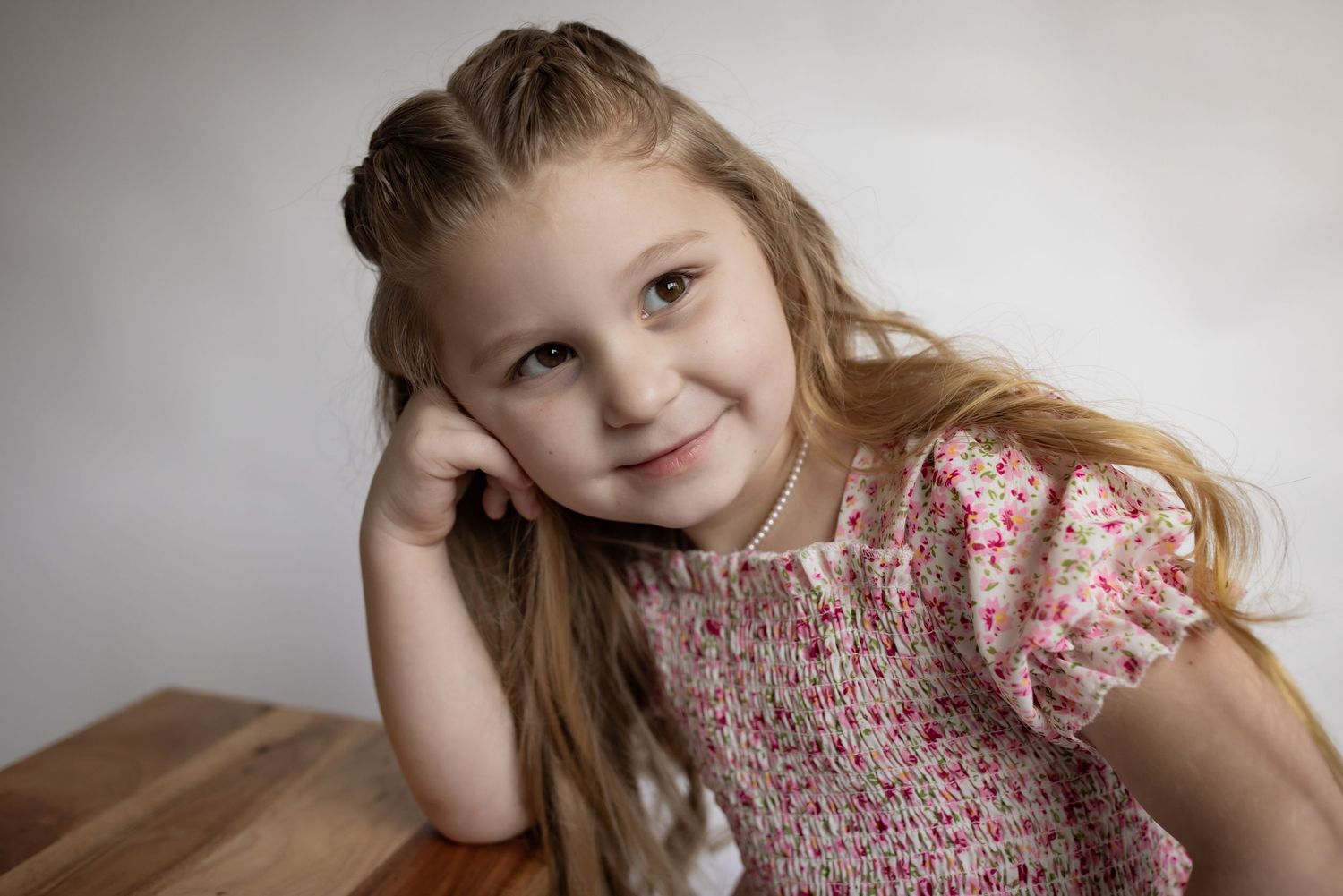 Young girl with long blonde hair in a floral dress resting her head on her hand at a wooden table.