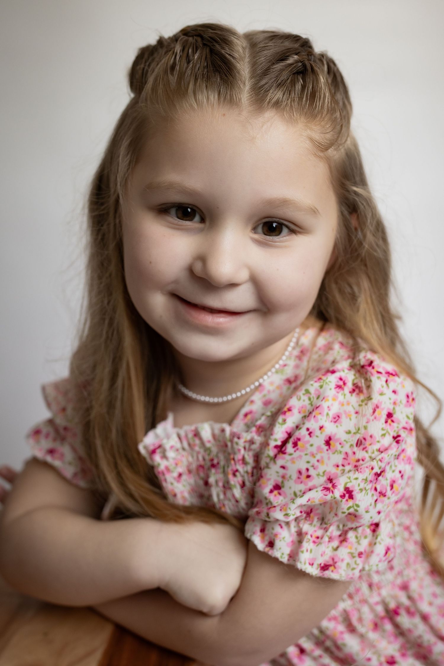 Young girl with long blonde hair in a floral pink dress and pearl necklace smiling with arms crossed.