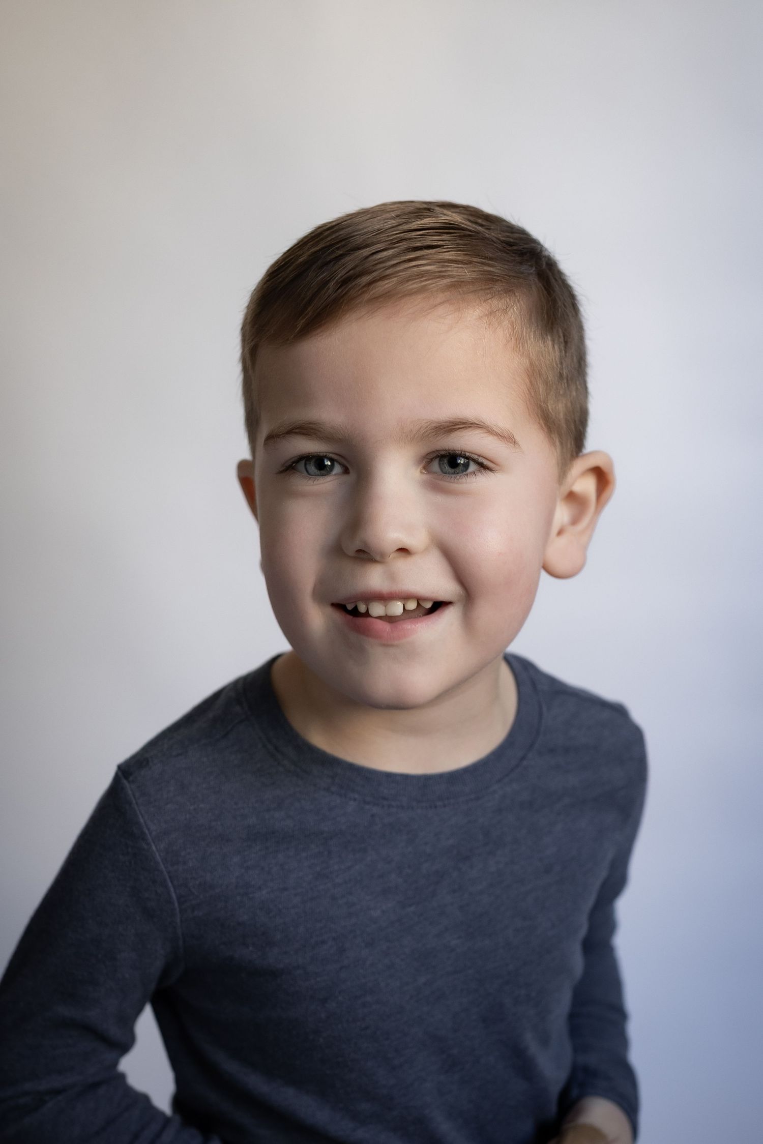 Happy young boy with short brown hair smiles wearing a grey long sleeve shirt.