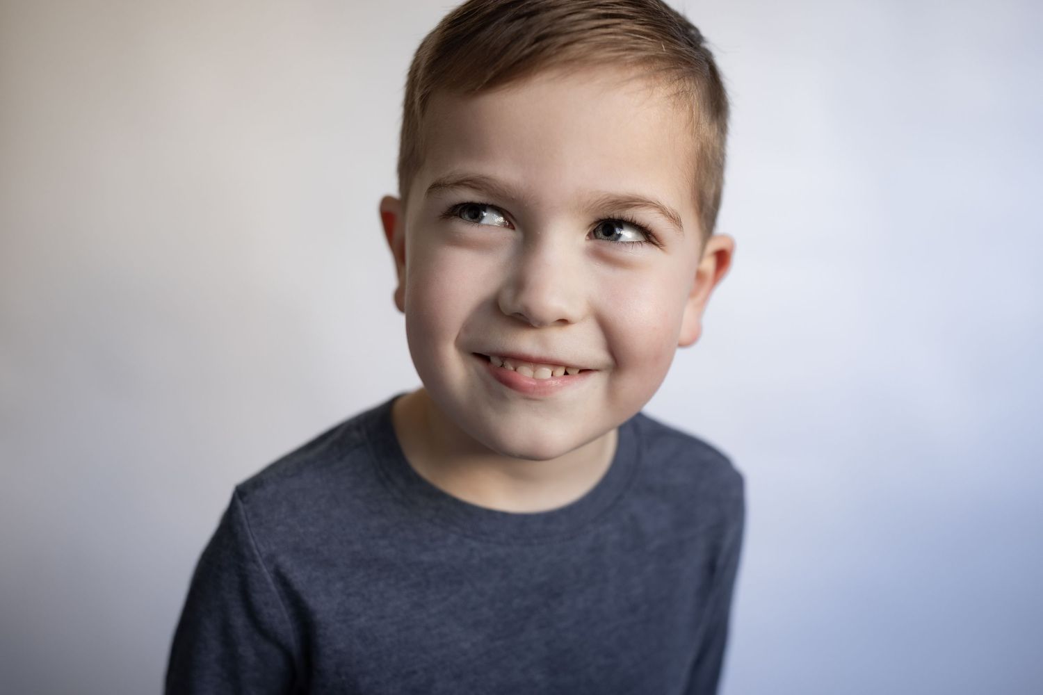 Young boy in grey long sleeve shirt smiles and glances sideways against white background.