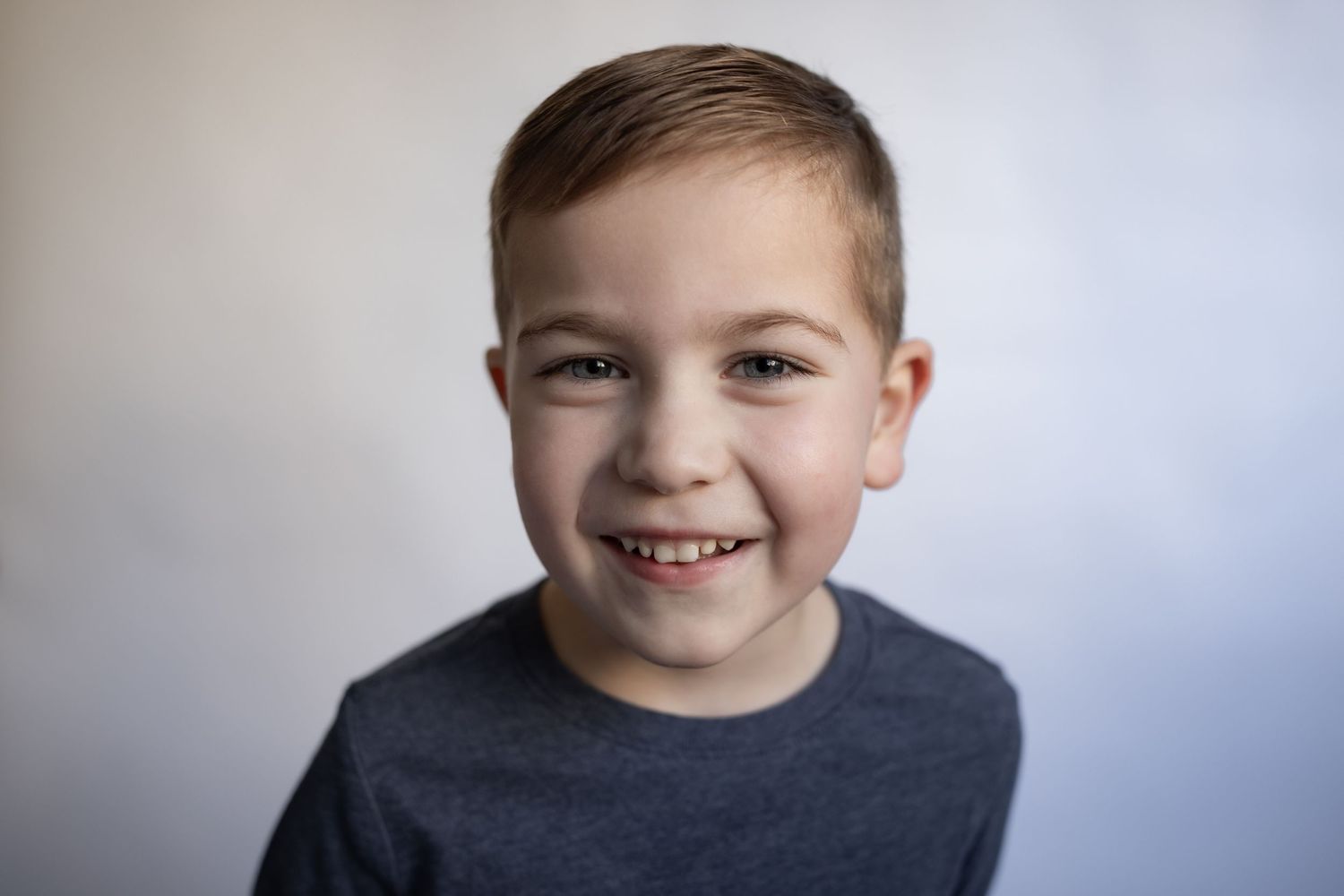 Smiling young boy with short brown hair wearing a gray t-shirt against a white background.