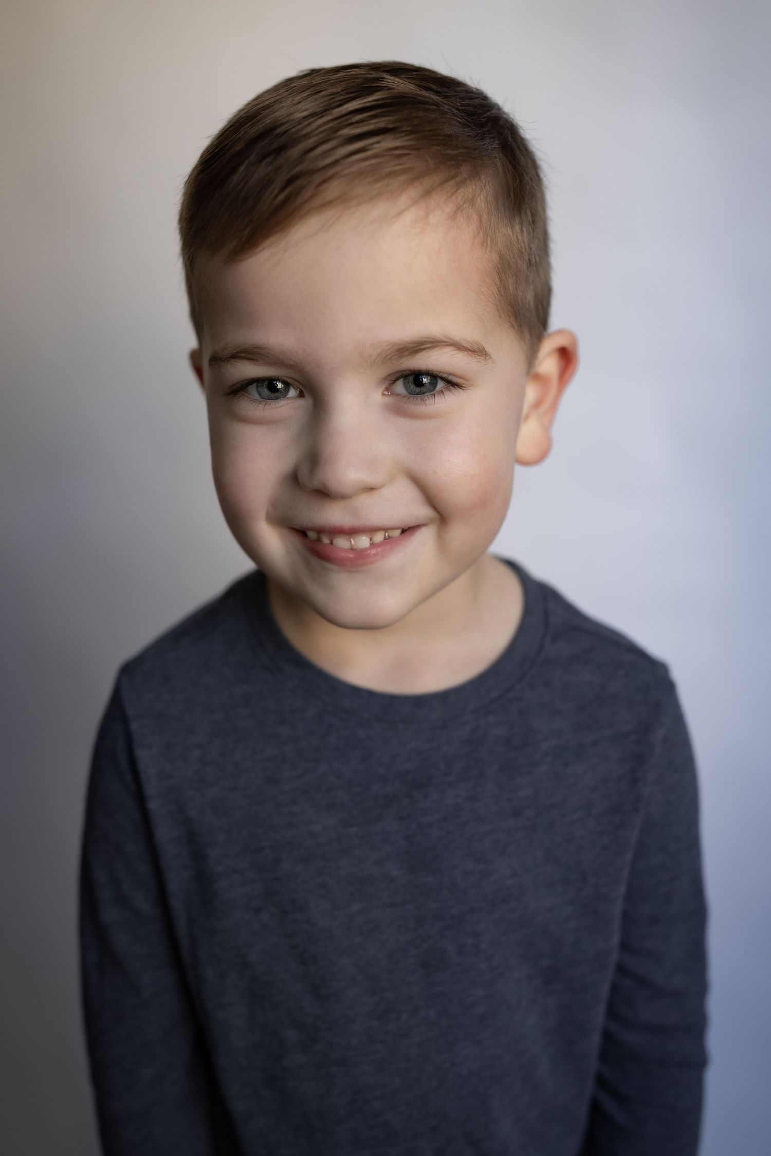 Cheerful young boy with neat brown hair smiles warmly wearing a heather grey shirt.