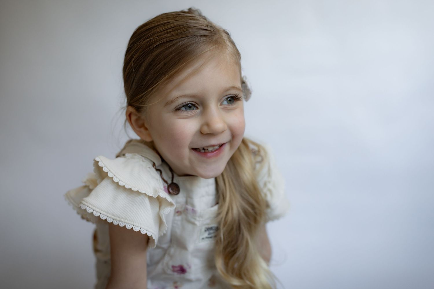 Smiling blonde girl in ruffled floral OshKosh top with side ponytail looks playfully.