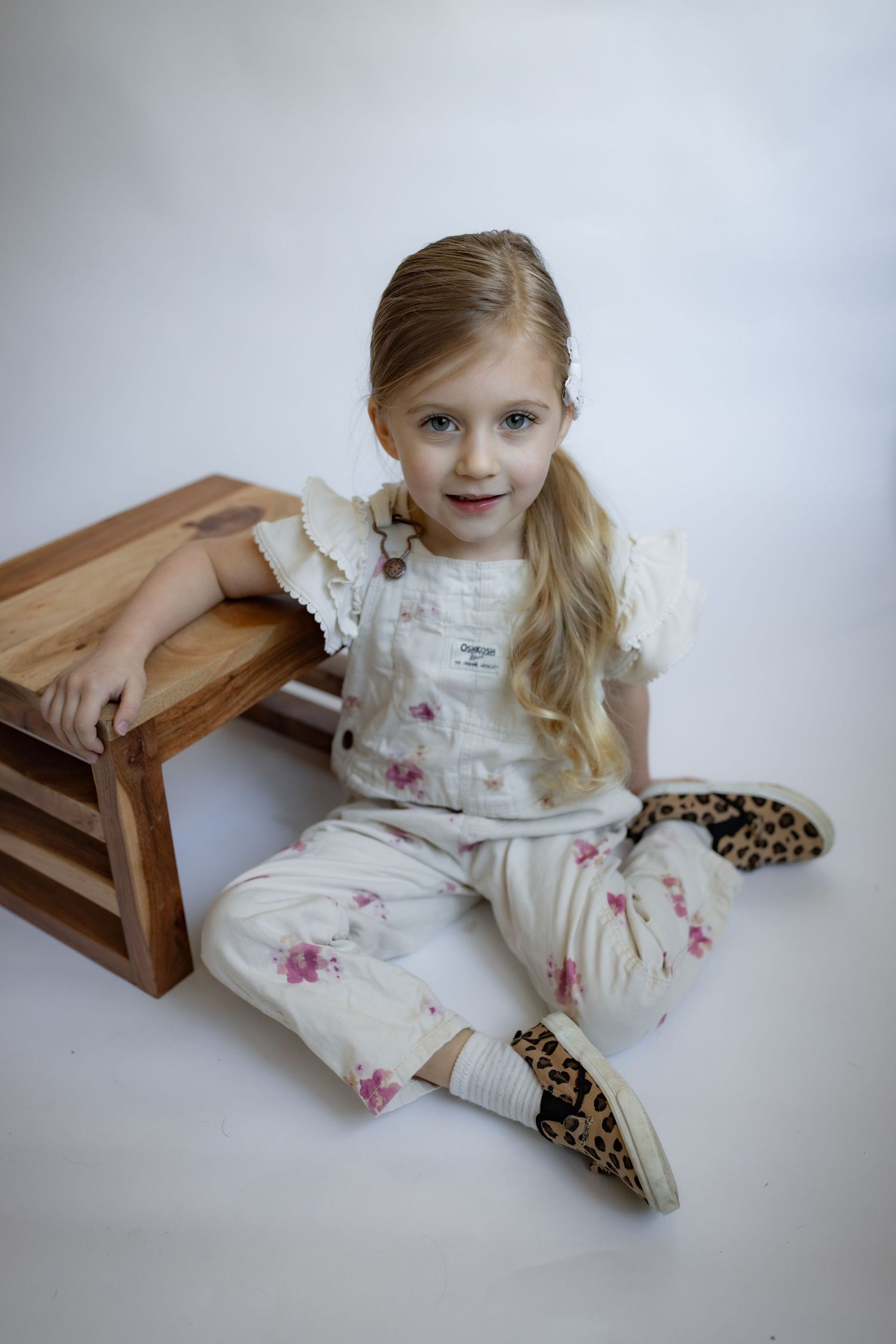 Blonde girl in floral overalls sits on floor beside wooden crate wearing leopard boots.
