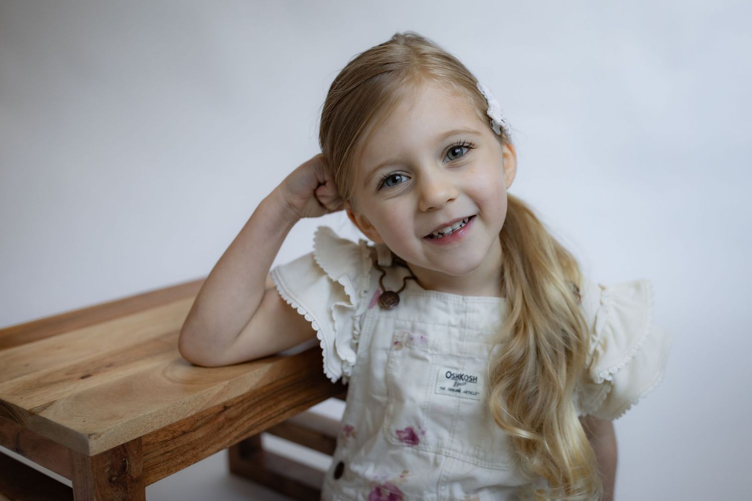 Cheerful blonde girl in floral OshKosh outfit leans on wooden bench smiling brightly.