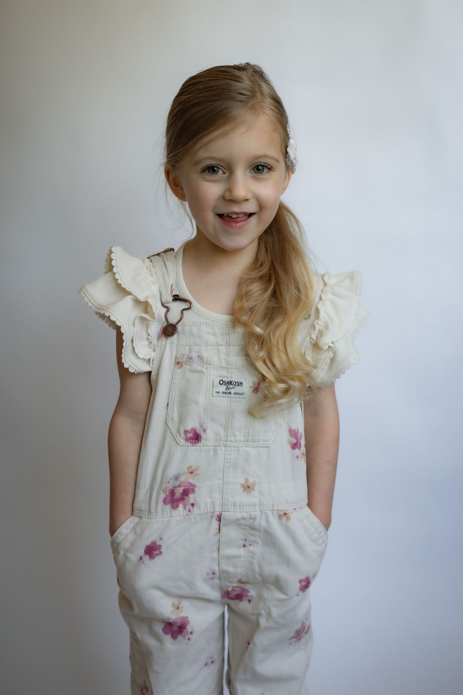 Smiling blonde girl in floral OshKosh overalls with ruffled cream top stands cheerfully.