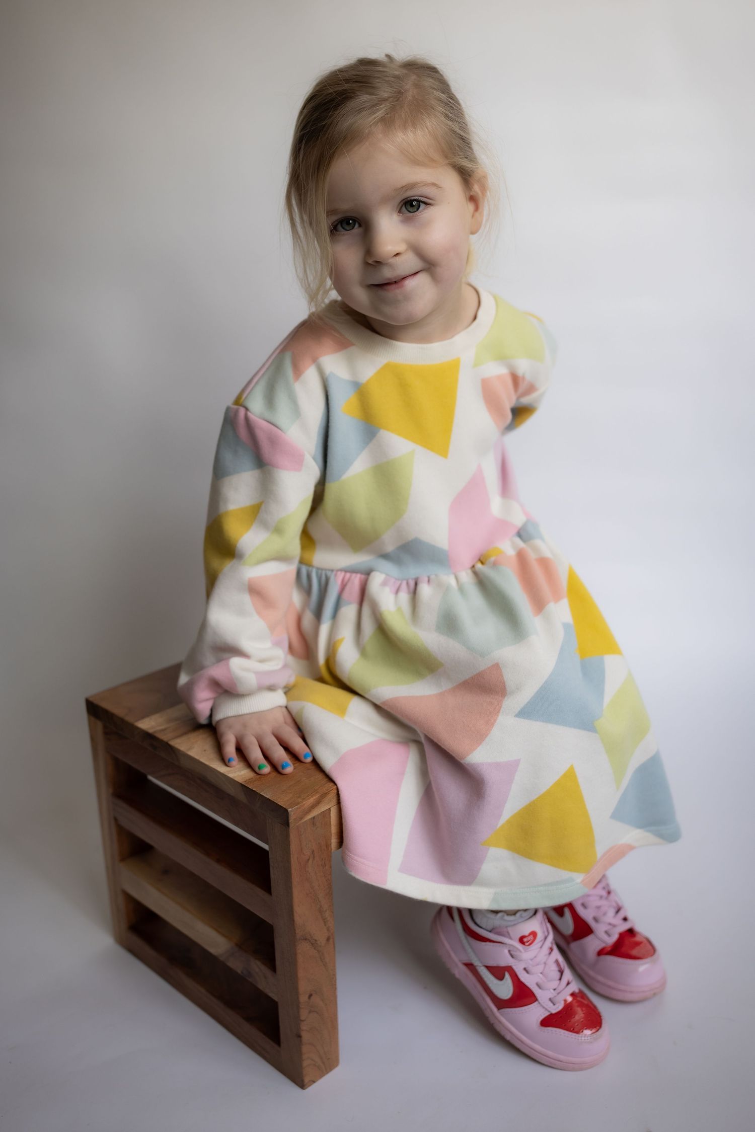 Young girl in colorful triangle-print dress and pink sneakers sitting on wooden crate.
