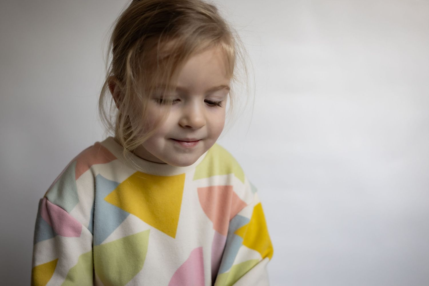 Cute toddler girl in colorful geometric triangle print sweatshirt looking down shyly.