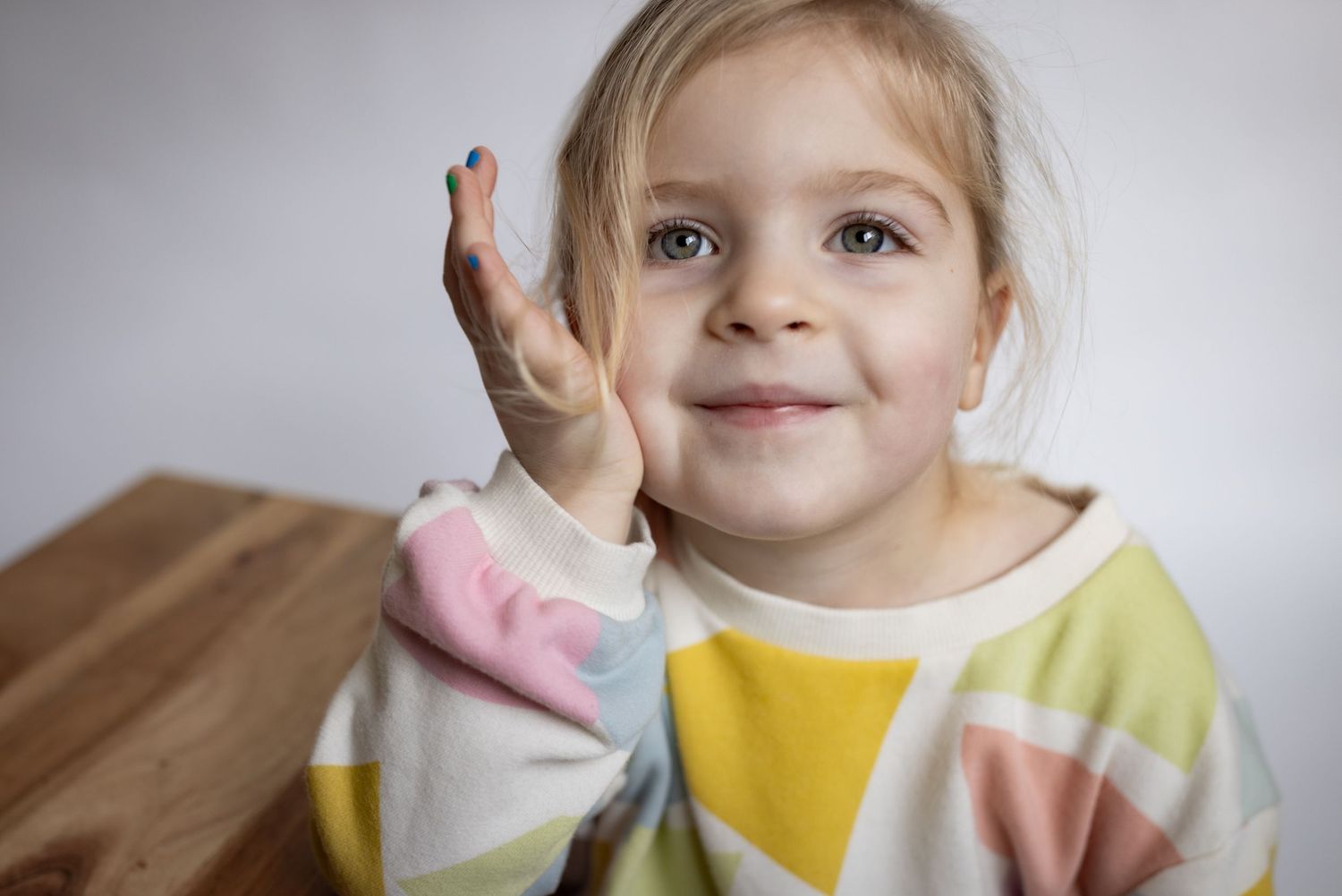 Playful blonde toddler girl in colorful geometric print top raising hand near face.