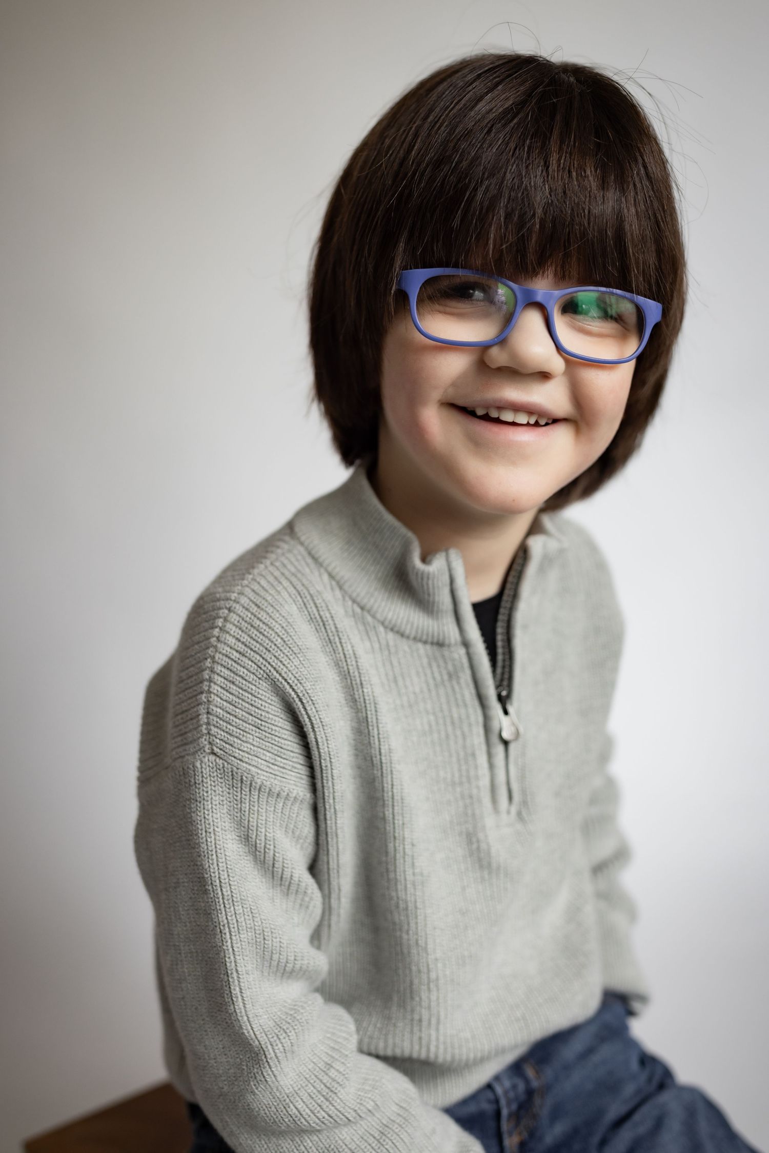 Smiling young boy with blue glasses and gray sweater posing for portrait photo.