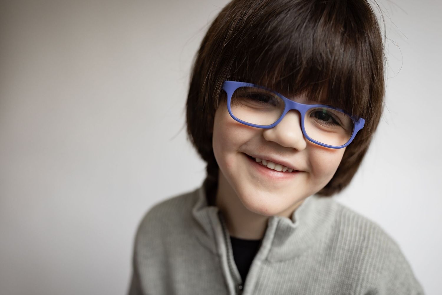 Smiling young boy with dark bowl-cut hair wearing blue-framed glasses and a gray zip-up jacket.