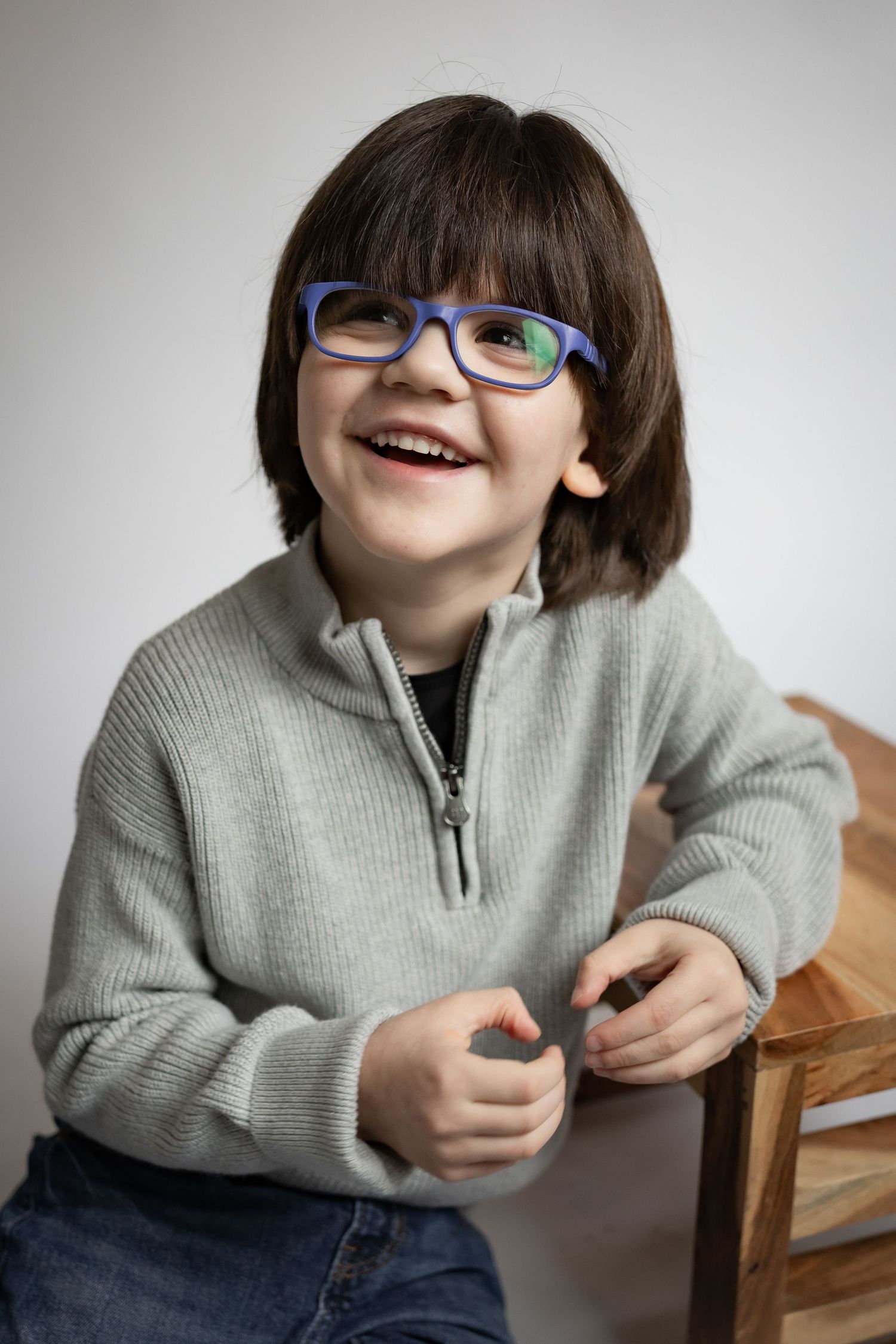 Happy young boy with blue glasses and gray sweater laughing while sitting on wooden stool.