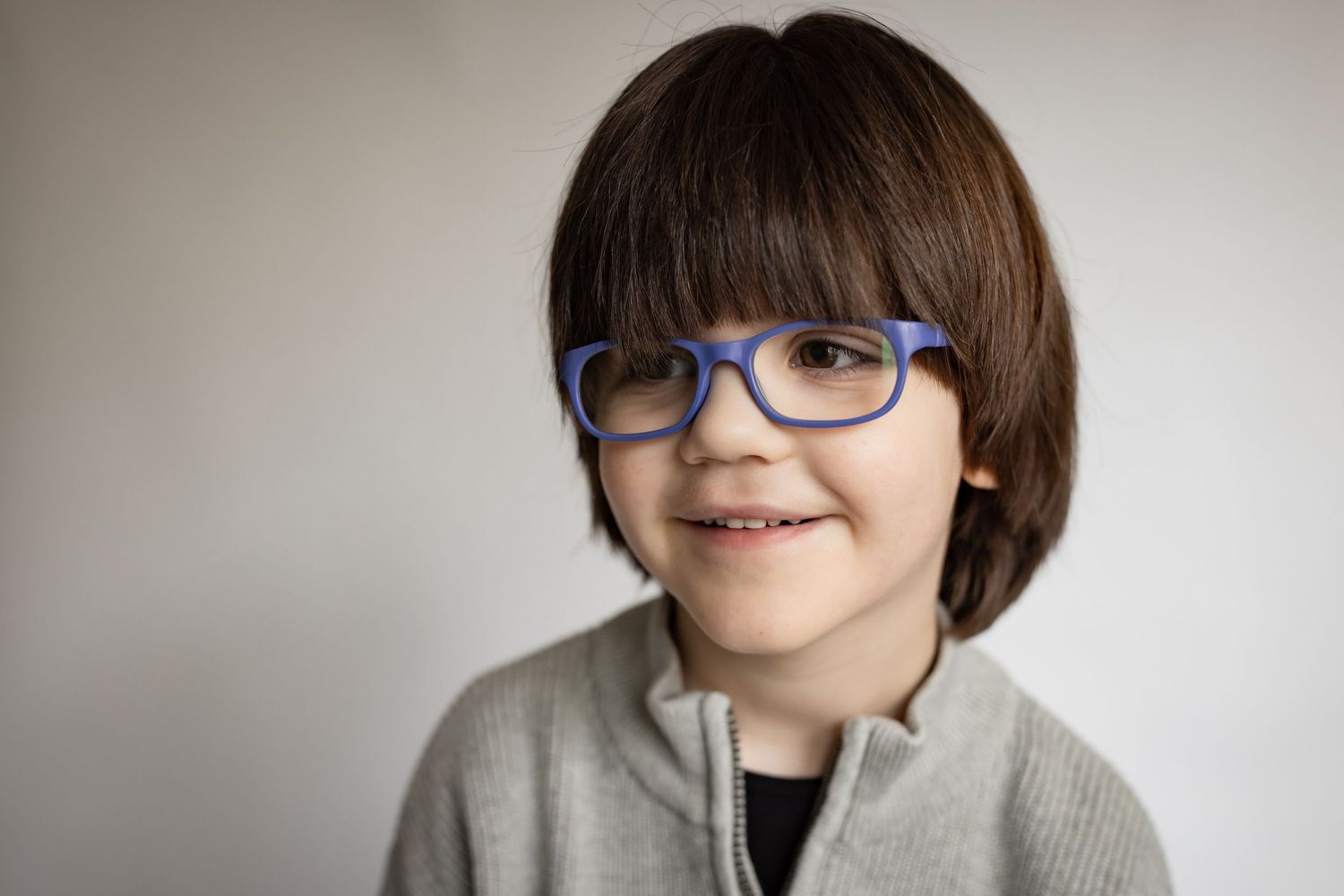 Cheerful young boy wearing blue rectangular glasses and gray zip-up sweater smiling.