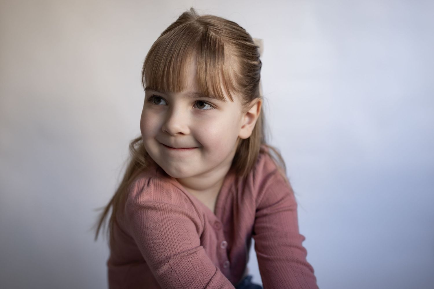 Cute young girl with bangs in pink ribbed top smiling softly at camera.