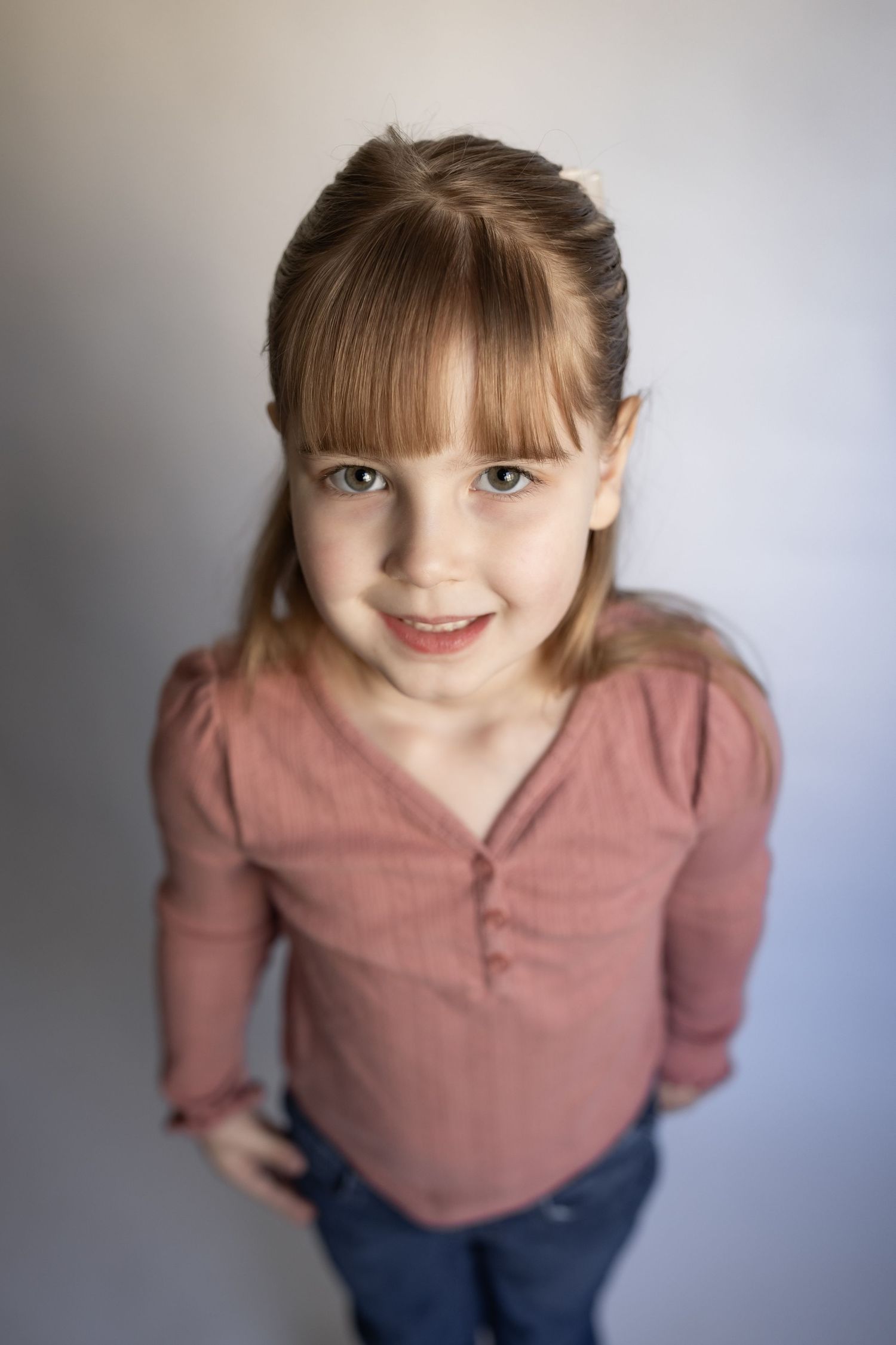 Young girl with bangs in pink henley top standing and smiling at camera.
