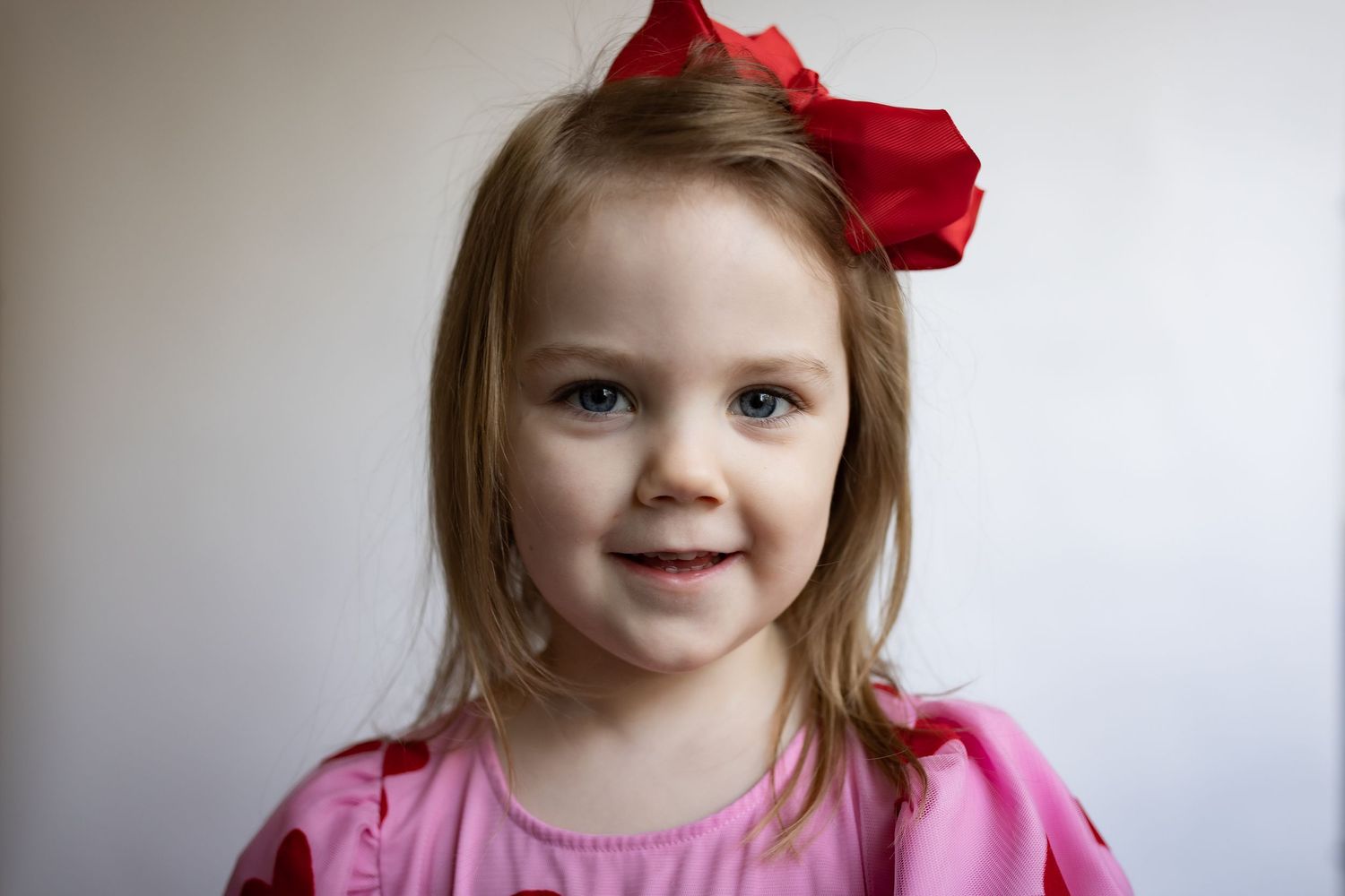 Cute young girl with blonde hair wearing a large red bow and pink dress, smiling at the camera.