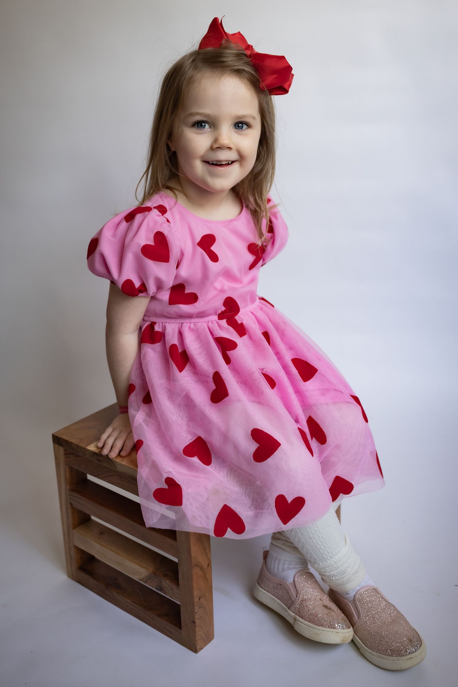 Adorable little girl in pink heart-print dress with red bow sitting on wooden crate.