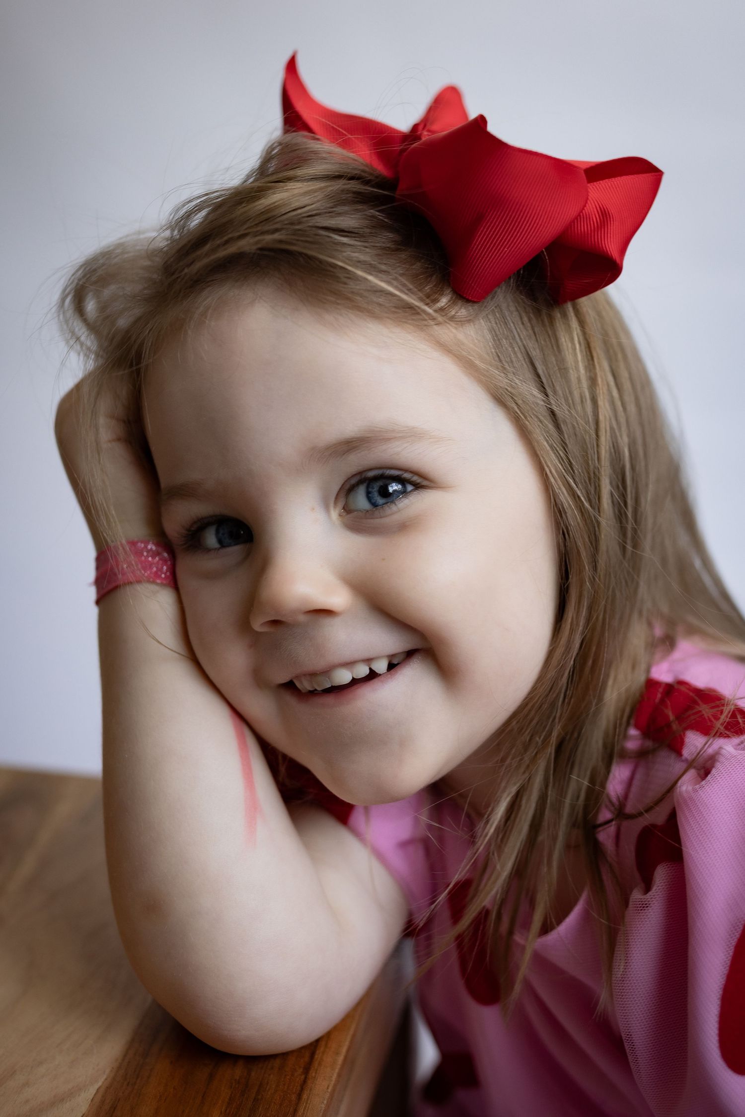 Sweet little girl with red bow hair accessory smiling with chin resting on hand.