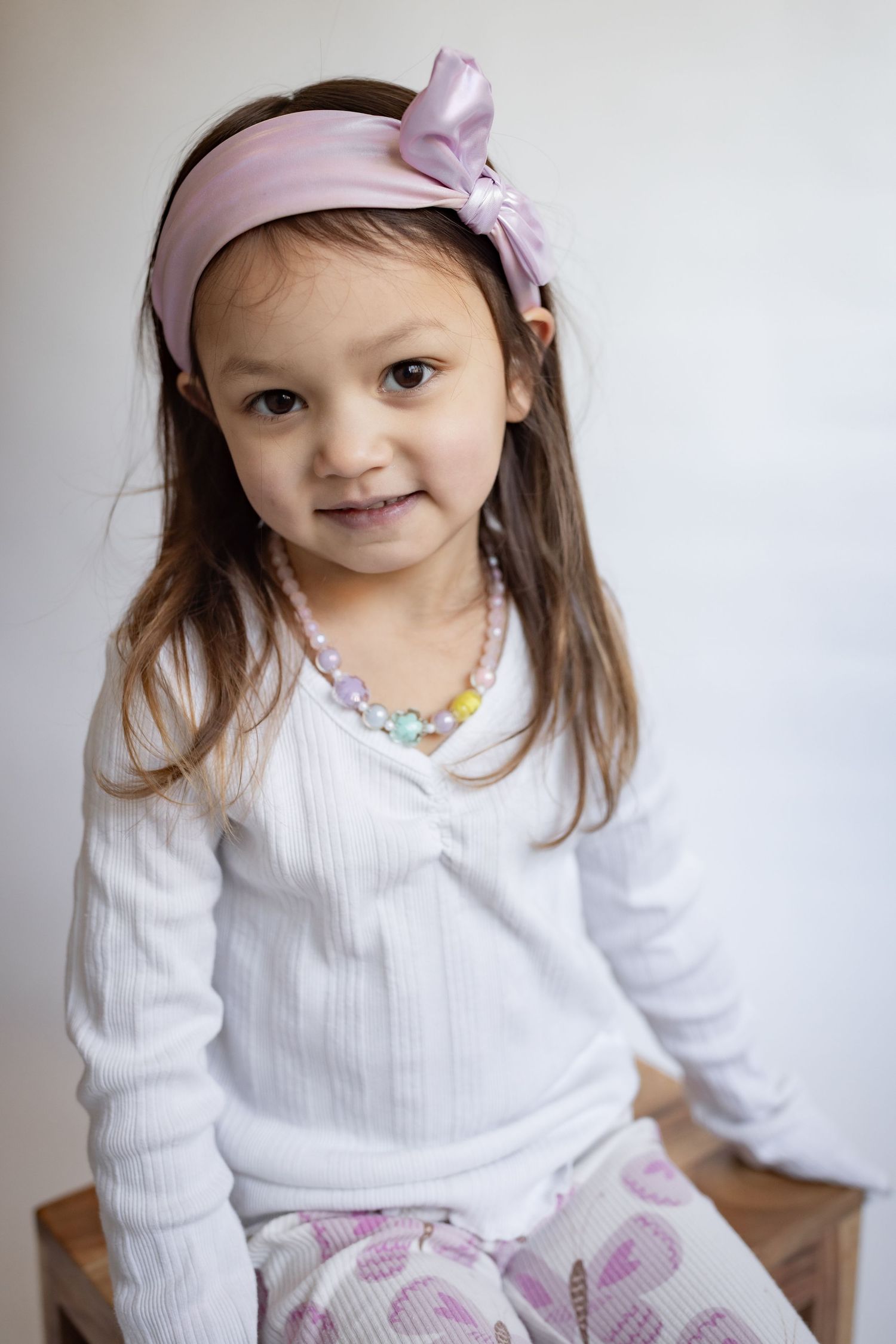 Adorable little girl wearing pink bow headband and colorful beaded necklace smiles softly.