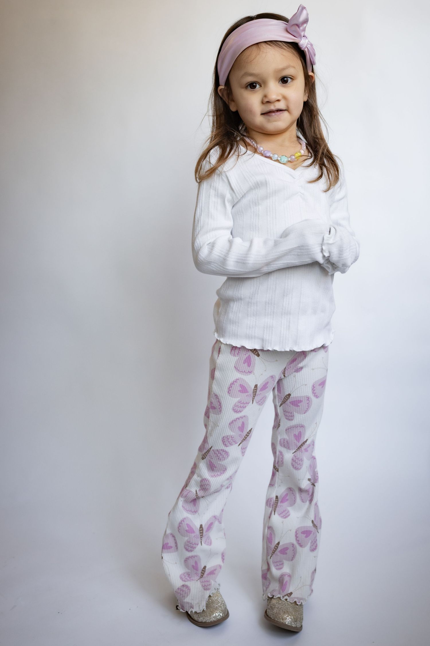 Young girl in white top and floral purple butterfly pants with pink headband poses confidently.