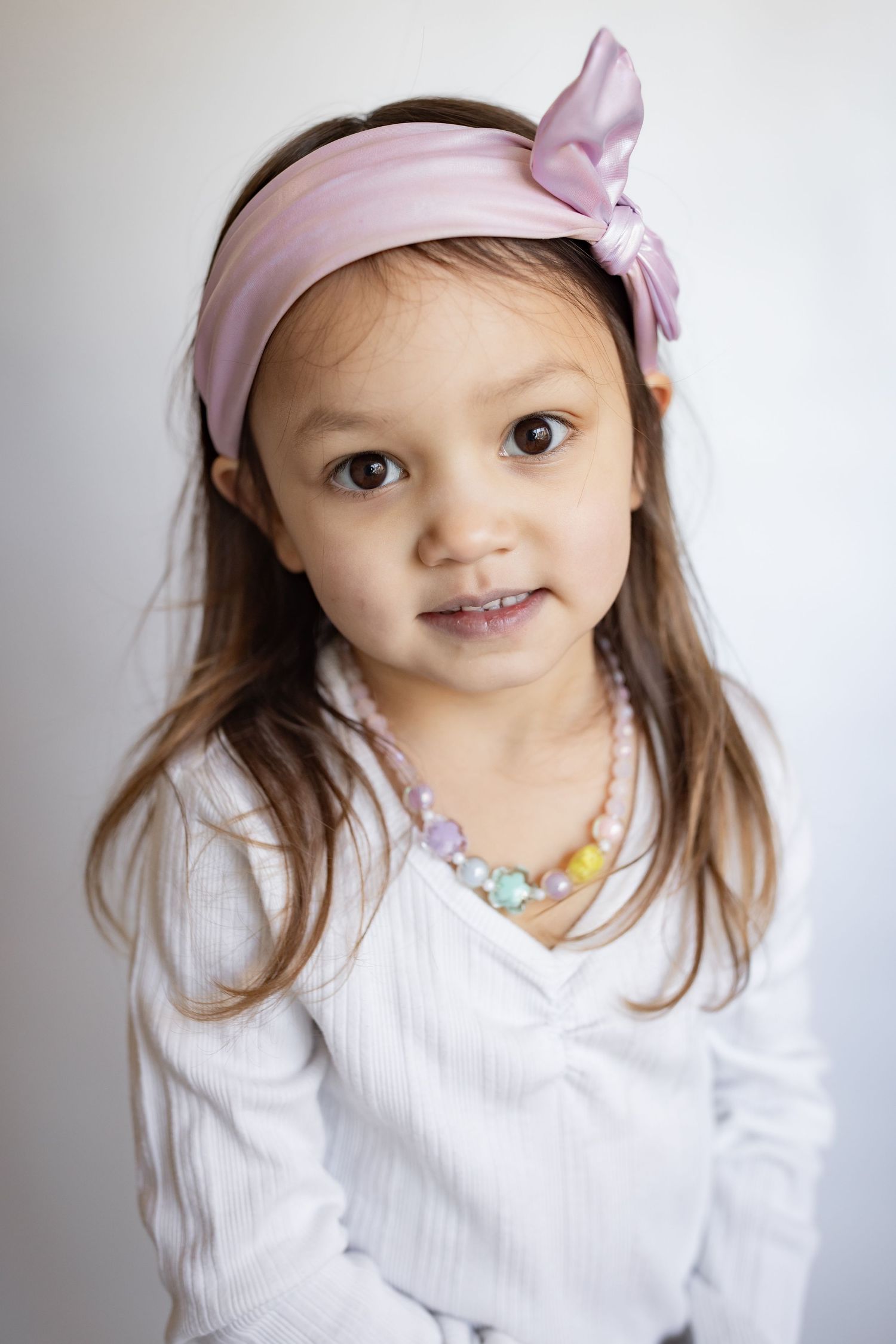 Adorable young girl wearing a pink bow headband, colorful bead necklace, and white top, smiling gently.