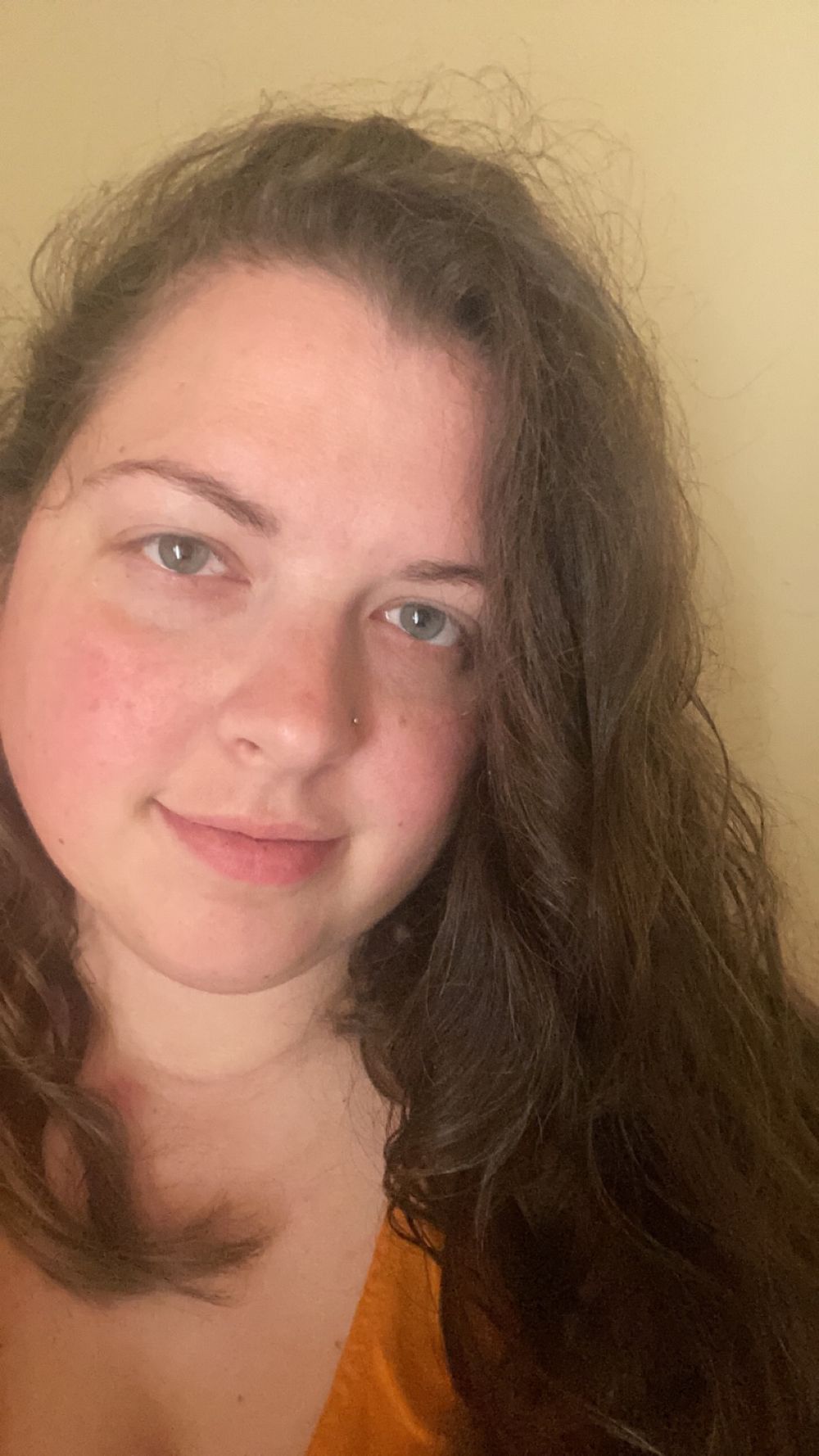 A selfie portrait showing long dark wavy hair and a warm smile against a neutral background, wearing an orange top.