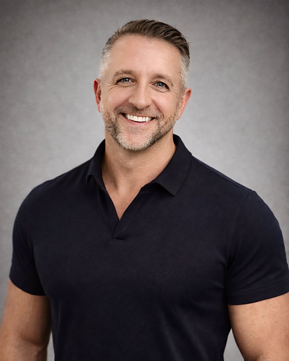 Studio portrait of person with gray hair and beard smiling warmly while wearing a black polo shirt.