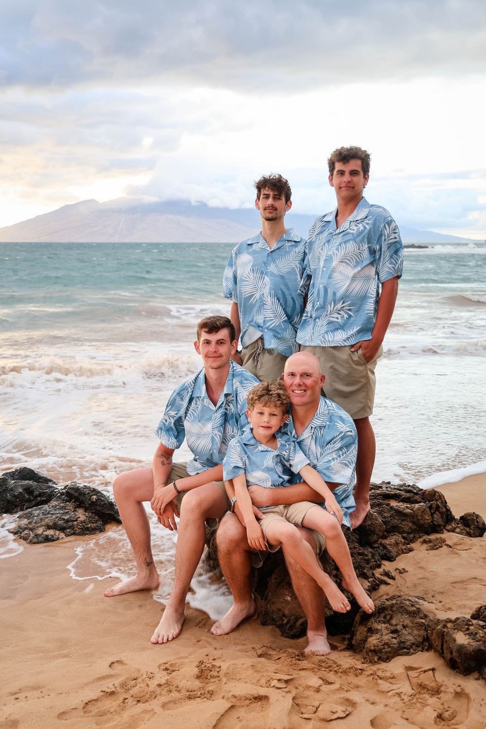 Family group wearing matching blue Hawaiian shirts pose together on rocky beach shore at sunset in Maui.