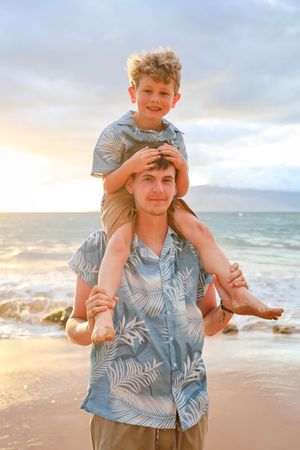 A parent and child play together on a sandy beach at sunset in matching blue palm print shirts.