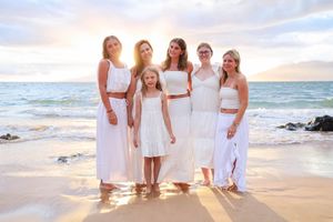A group in white dresses stands together on a sandy beach at sunset with ocean waves in the background.