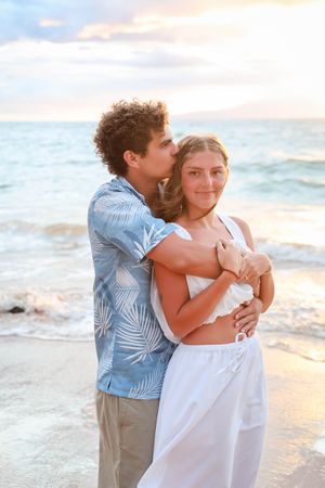 A couple embraces on a sandy beach during golden hour with the ocean and sunset creating a romantic backdrop.