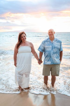 A couple stands together on a beach at sunset with ocean waves washing over their feet in coordinating blue and white outfits.