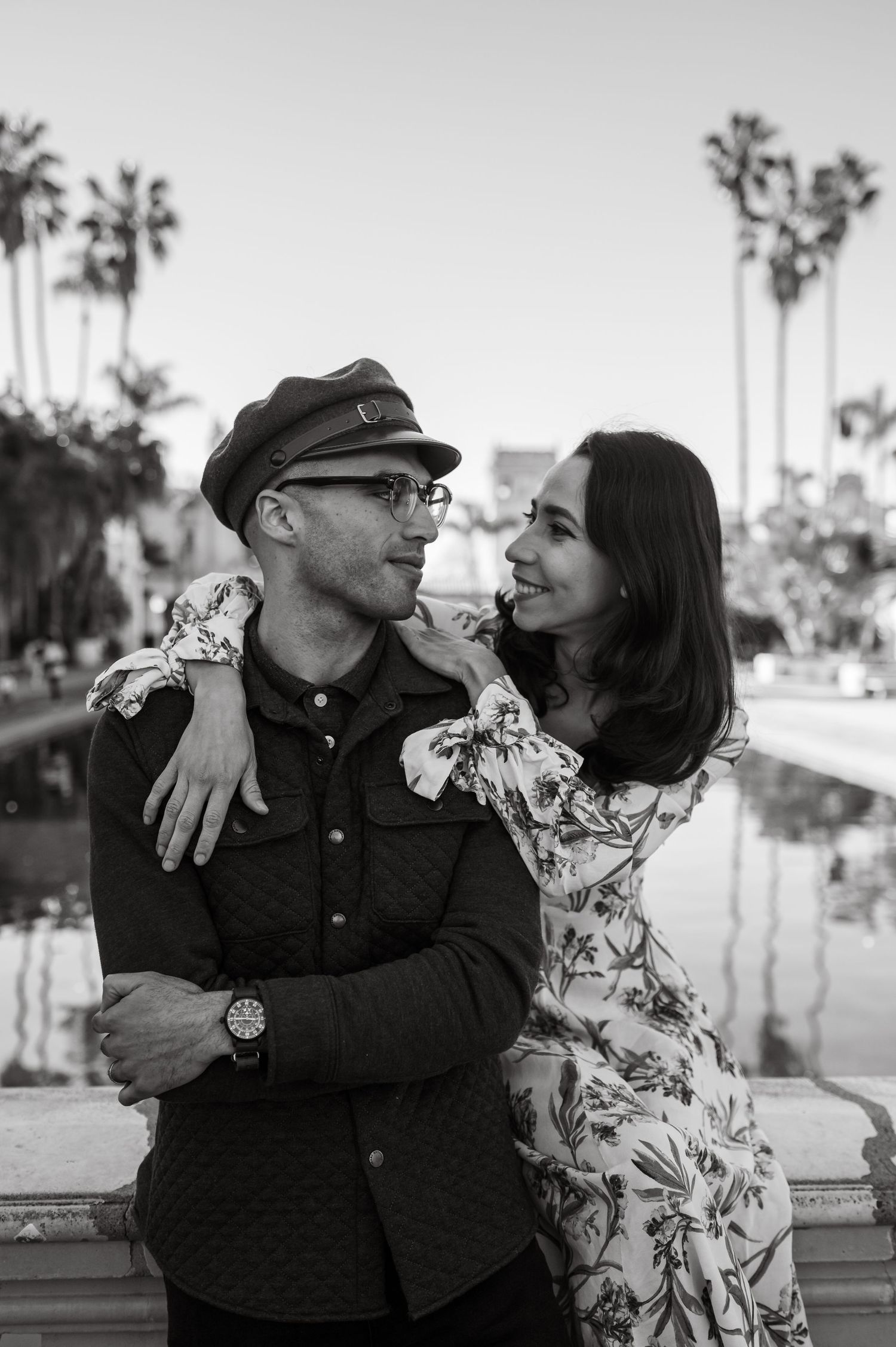 A couple poses against a concrete wall, wearing contrasting outfits of a floral dress and dark button-down shirt.