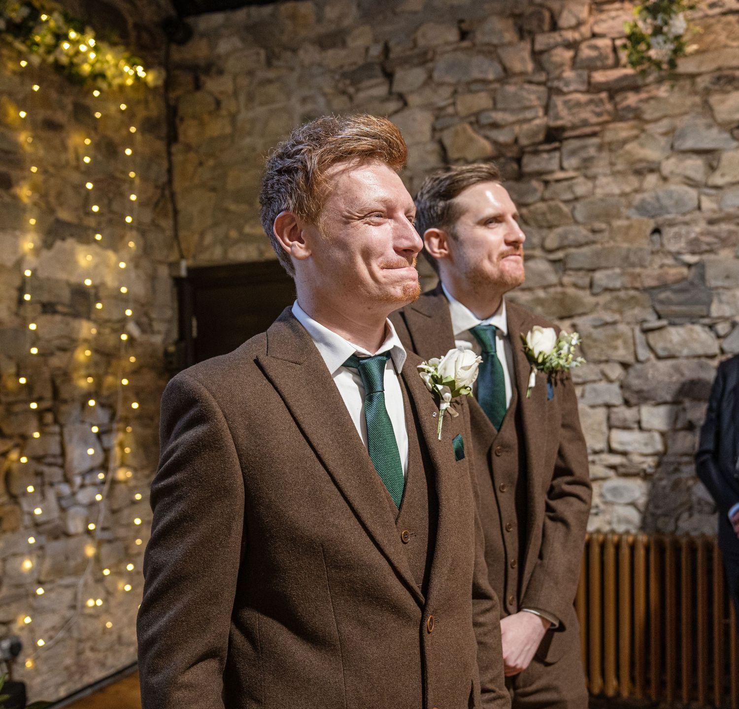 Groomsmen in brown suits and green ties laugh together against a rustic stone wall with twinkling lights.