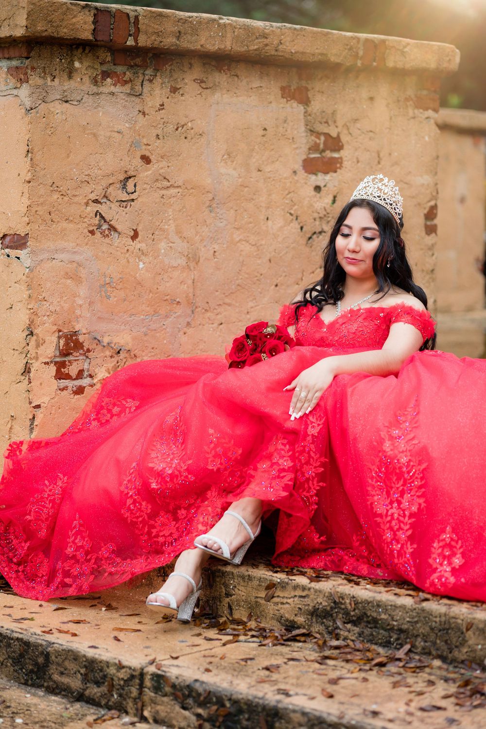 Young woman in red quinceañera ball gown with crown resting on stone steps.