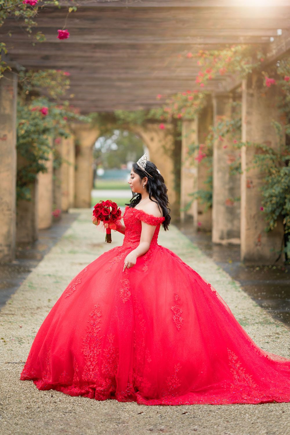 Quinceañera in stunning red lace ball gown holding bouquet under floral pergola.