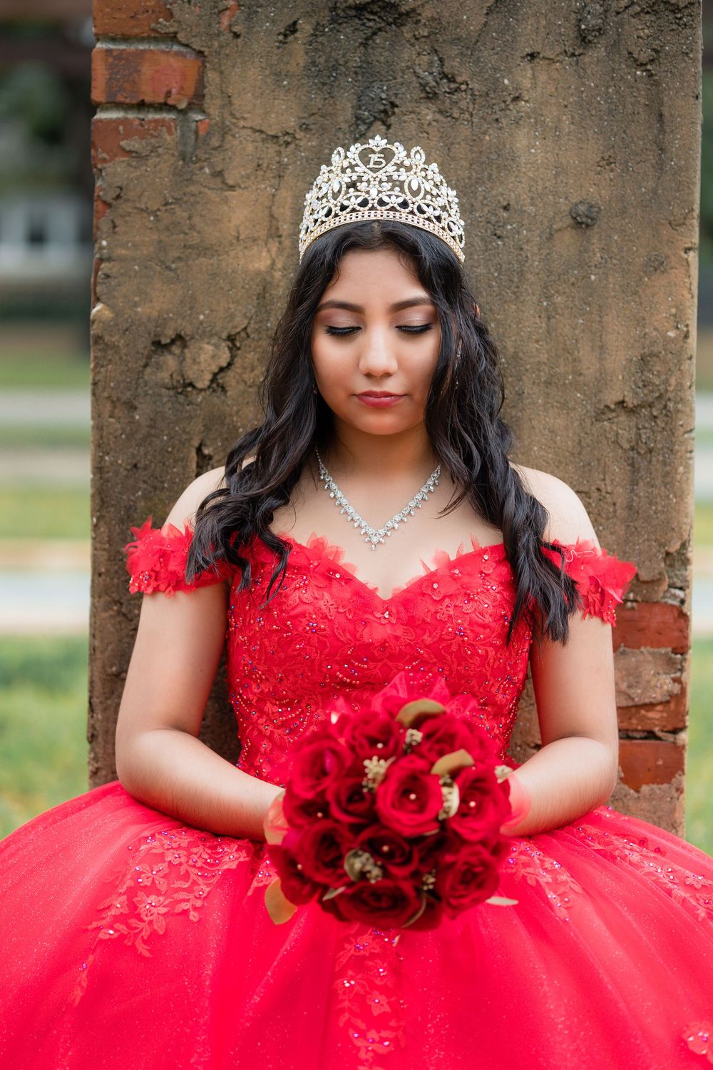Young woman in red quinceañera dress and crown holding red rose bouquet outdoors.