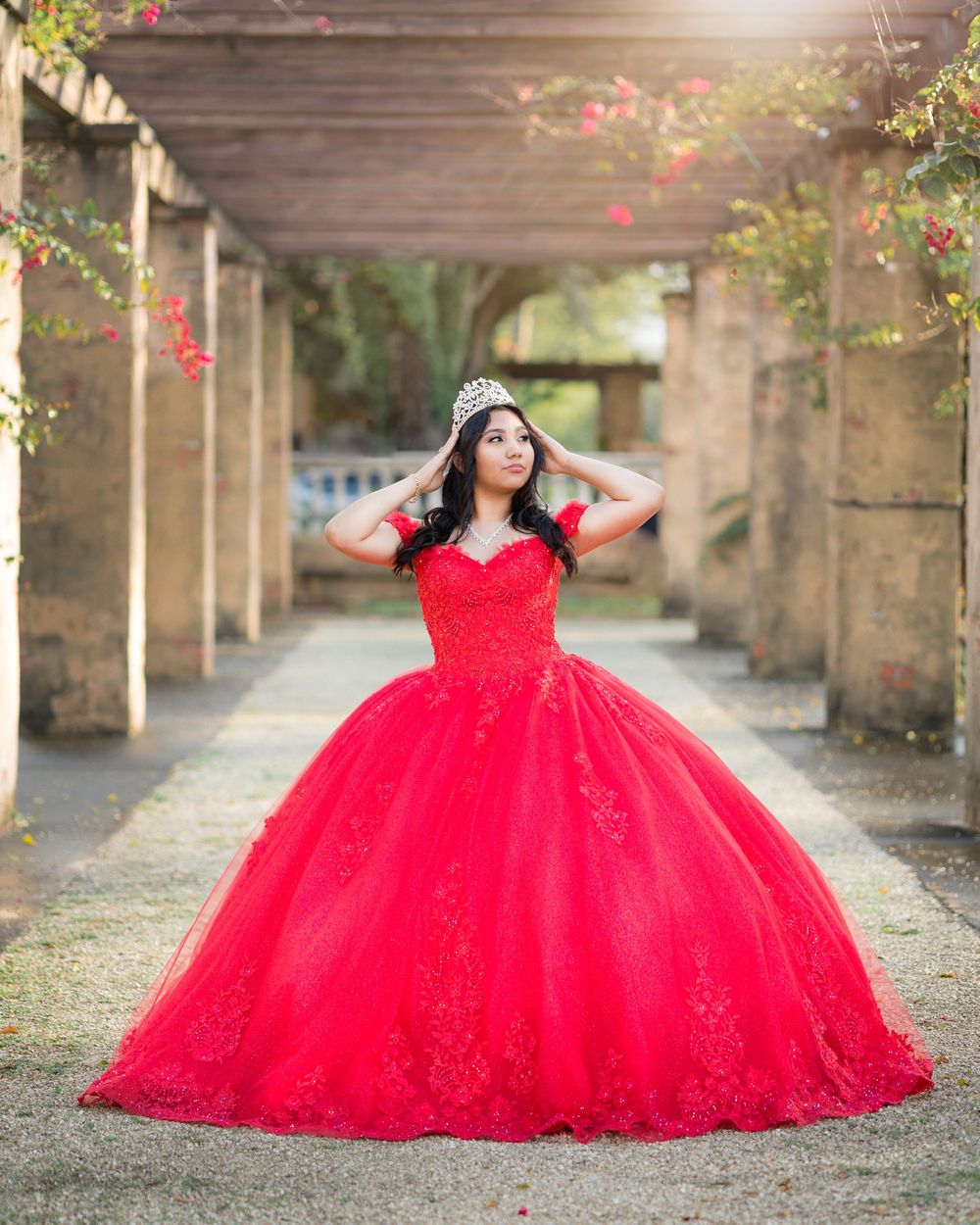 Quinceañera posing in voluminous red lace gown and tiara under garden pergola.