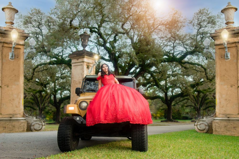Quinceañera in red ball gown posing on lifted truck at grand estate entrance.