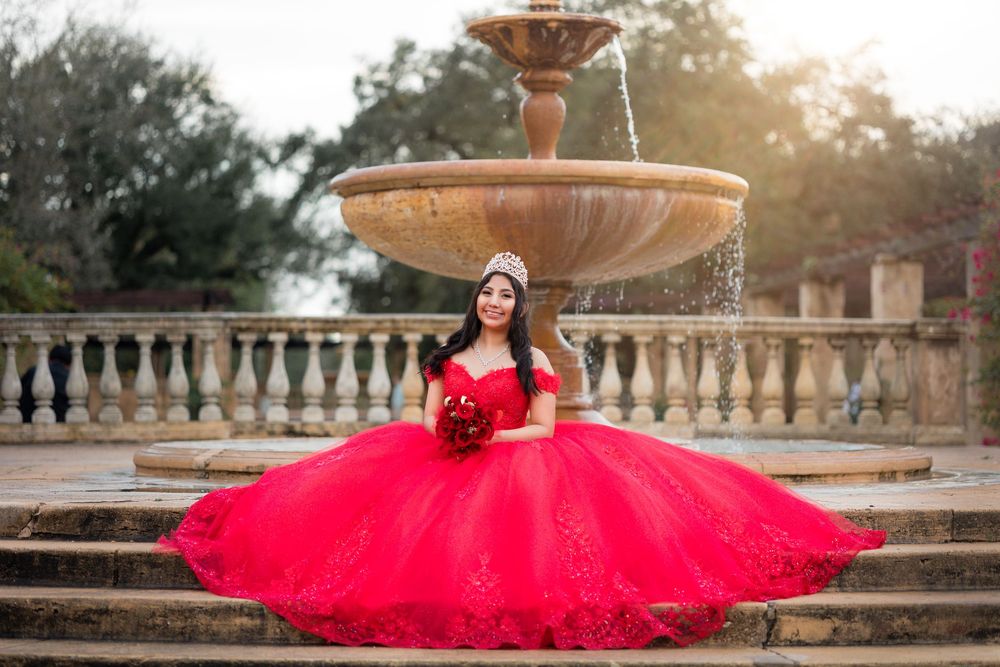 Young woman in red quinceañera ball gown with tiara poses on stone steps by ornate fountain.